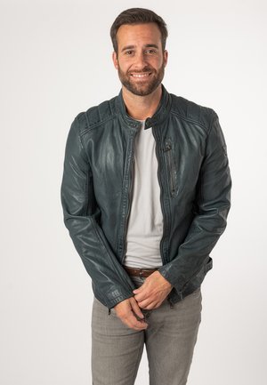 Donker turkooise leren jas met een ritssluiting aan de voorkant, een bandkraag en gestikte schouderdetails. Draagt over een lichtgrijze shirt.