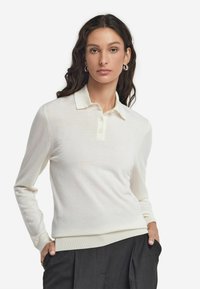 Långärmad polo-tröja i mjuk krämfärgad tyg med krage, tre franska knappar och en åtsittande fåll, kombinerad med mörka byxor.