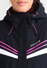 Námořnická zimní bunda s růžovými a bílými šikmými pruhy, nastavitelnou kapucí a logem viditelným na hrudi; vyrobená z materiálu odolného vůči povětrnostním vlivům.