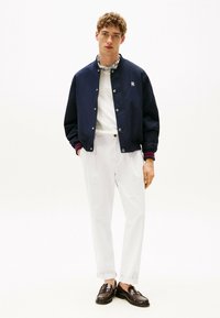 Giacca bomber navy con bottoni a pressione, felpa bianca, pantaloni chiari e mocassini scuri. Presenta polsini a coste con un accento bordeaux.