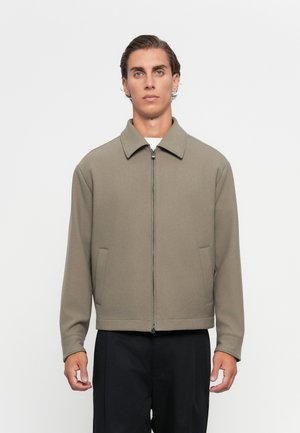 Olivgrön zip-up jacka med spridd krage, sidofickor och avslappnad passform. Materialet verkar strukturerat och mjukt.
