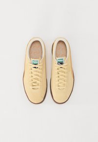 Baskets en daim beige avec des lacets assortis et une semelle en caoutchouc marron. Étiquettes vertes présentant le logo Puma sur la languette, avec des semelles intérieures à motifs.