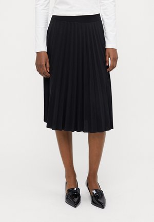 Kvinde iført en sort plisseret midi-nederdel, hvid langærmet top og sorte laklæder loafers med spidse tæer.