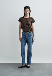 Brązowy krótki rękaw koszulki z nadrukiem panterki, niebieskie spodnie typu straight-leg z ozdobami oraz czarne płaskie buty. Prosty, codzienny strój.