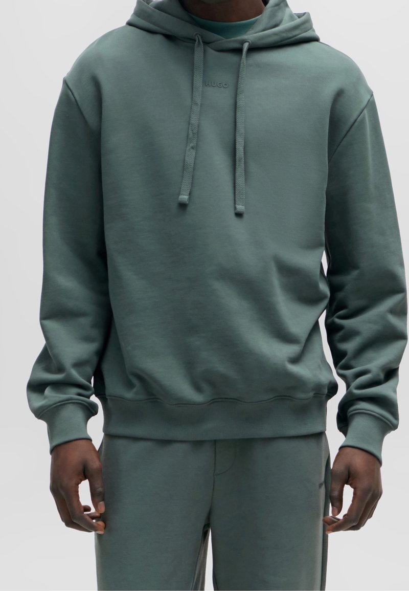 Homme portant un sweat à capuche vert atténué avec des cordons et un pantalon assorti, debout devant un fond clair uni.