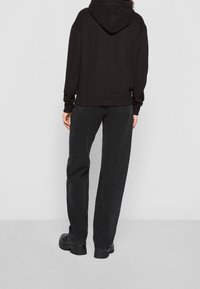 Sudadera negra con capucha y corte holgado, combinada con vaqueros negros de talle alto y zapatos negros gruesos, sobre un fondo liso.