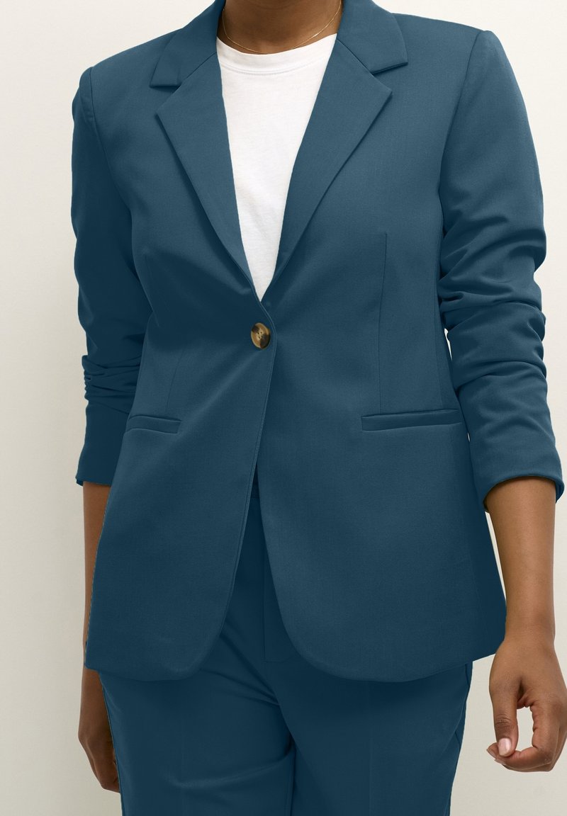Personne portant un blazer ajusté bleu sarcelle avec un seul bouton marron sur un t-shirt blanc à col rond, avec un pantalon bleu sarcelle assorti.