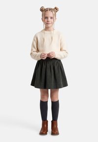 Jeune fille debout portant un pull crème, une jupe en velours côtelé foncé, des chaussettes grises hautes jusqu'aux genoux, des bottines marron, avec les cheveux tressés en deux chignons.