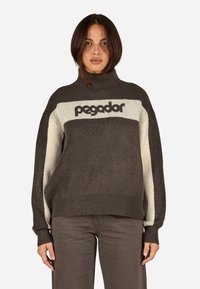 Tamsiai rudas mezginiu su aukštu kaklu, su grietinės spalvos juosta ir žodžiu "pegador" tamsiomis raidėmis krūtinėje.