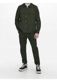 Donkergroene collared jas met twee voorkruiken, gecombineerd met bijpassende broeken. Zwarte sneakers met een gestructureerde zool maken de outfit compleet.