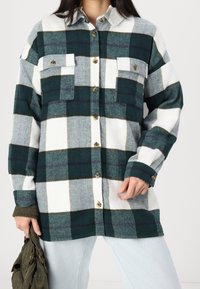 Koszula flanelowa w kratę w kolorach zielonym, białym i szarym. Posiada dwie przednie kieszenie, guziki oraz długie rękawy. Miękka w dotyku i o luźnym kroju.