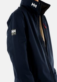 Chaqueta impermeable de color navy con un alto cuello, presenta el logo blanco de Helly Hansen y un tirador de cremallera rojo. Textura suave, tejido ligero.