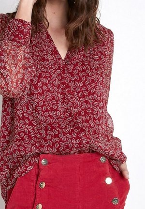 Vrouw die een doorschijnende rode blouse met lange mouwen en een wit bladmotief draagt, en een hoge taille rode corduroy rok met zes metalen knopen.
