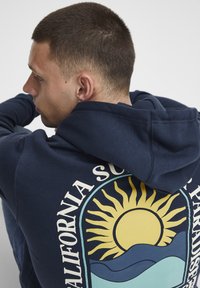 Marineblå hettegenser med et grafisk design av en sol og bølger i gult og teal på baksiden, laget av mykt materiale og løs passform.
