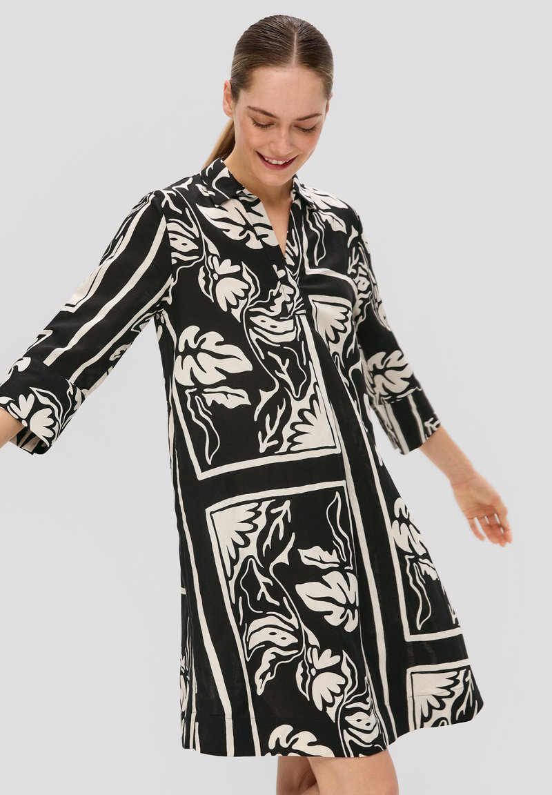Robe florale noir et blanc avec des manches longues, un col, et une coupe ample, présentant des rayures audacieuses et différents motifs de feuilles.