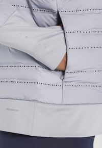 Ręka wsunięta w boczną kieszeń jasnoszarej kurtki Nike Aeroloft z perforacjami, z poziomymi czarnymi kropkami.