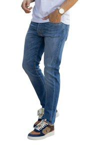 Man die blauwe slim-fit jeans, een wit overhemd, een horloge aan de linkerpols en meerkleurige sneakers met witte veters draagt, staand met één hand in zijn zak.