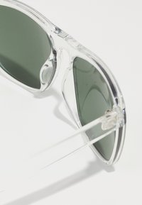 Ochelari de soare dintr-un plastic transparent, cu lentile verzi, având un design rotunjit și detaliu la balama. Cadru ușor cu o textură netedă.