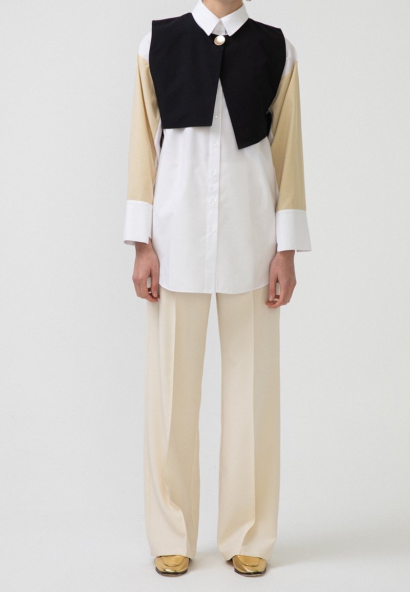 Personne portant un pantalon beige à jambes larges, une longue chemise beige et blanche, un gilet court noir avec un bouton doré et des chaussures dorées à bout carré.