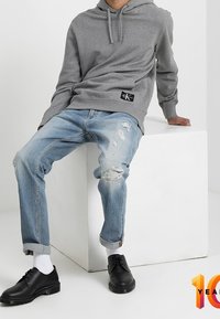Šedá mikina s kapucí a logem, spárovaná se světle modrými roztrhanými džíny a černými šněrovacími botami. Bílé ponožky viditelné.