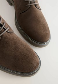 Chaussures en daim marron avec une texture lisse, bout rond et coutures contrastantes le long de la semelle. Présentent des lacets marron et un talon robuste.