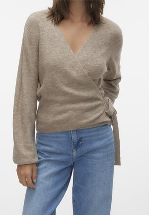 Kvinde iført en beige slå-om sweater med sidebind og lange ærmer, kombineret med blå denimjeans mod en ensfarvet baggrund.