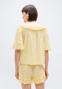 Femme aux cheveux courts bruns, portant un haut à manches courtes jaune pâle et un short assorti avec une bordure en dentelle festonnée, vue de dos.