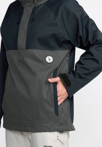 Veste à enfiler en bleu foncé et gris, dotée d'une demi-fermeture éclair, d'un écusson logo, d'une poche avec Velcro et d'un tissu lisse et résistant aux intempéries.