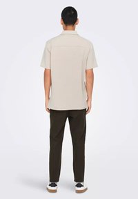 Camisa corta de color beige claro con cuello clásico, dos costuras en la parte trasera, combinada con pantalones verdes oscuros y zapatillas blancas con suelas marrones.