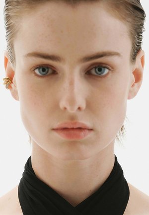 Pendiente de oro en la oreja con tres espirales entrelazadas, llevado en la oreja izquierda, sobre un fondo neutral. Piel suave con un acabado ligero de maquillaje.