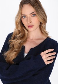 Blonde vrouw met blauwe ogen draagt een donkerblauwe fuzzy trui, met gekruiste armen en haar schouder tegen een effen lichte achtergrond.