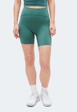 Mujer con pantalones cortos deportivos ajustados verdes, top corto a juego, calcetines blancos y zapatillas blancas para correr, de pie sobre un fondo liso.