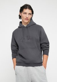 Sudadera gris oscura con un bolsillo canguro en el frente, capucha con cordones y logotipo en relieve de "Originals". La tela parece suave y texturizada.