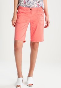 Shorts corail en tissu léger avec une coupe droite, poches avant et fermeture par bouton, associés à des mules ouvertes blanches.