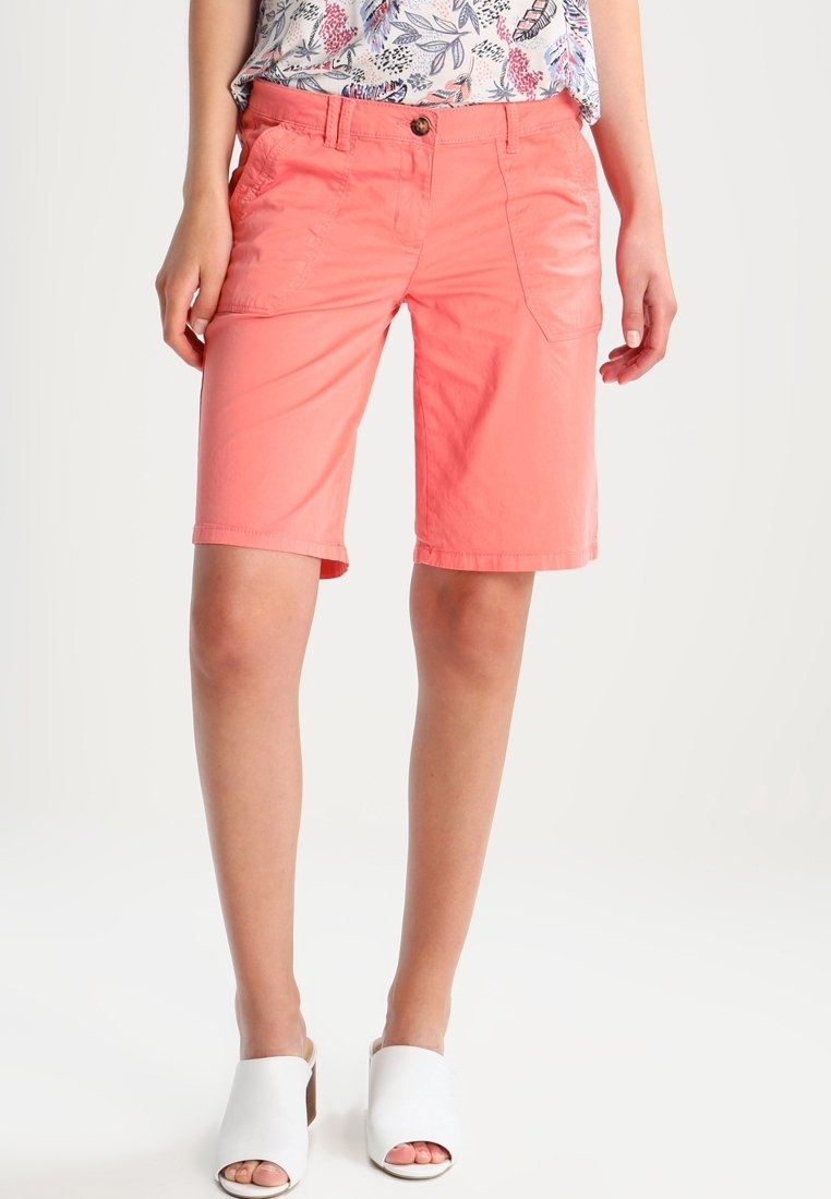 Shorts corail en tissu léger avec une coupe droite, poches avant et fermeture par bouton, associés à des mules ouvertes blanches.