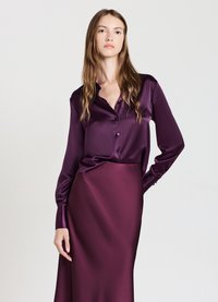 Camicetta in satin viola con chiusura a bottoni e maniche lunghe, abbinata a una gonna in satin viola coordinata, caratterizzata da una texture liscia e lucida.