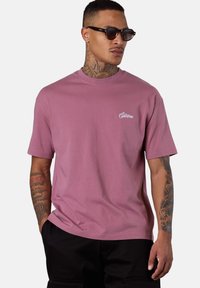 Mann mit Tattoos, der dunkle Sonnenbrillen, ein mauve T-Shirt mit kurzen Ärmeln und kleinem weißen Logo sowie schwarze Hosen trägt, steht vor einem einfarbigen Hintergrund.