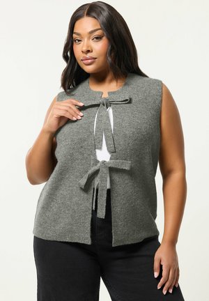 Frau trägt ärmellose graue Strickweste mit zwei vorderen Schleifen über weißem Oberteil und schwarzer Hose, posiert vor einem schlichten hellen Hintergrund.