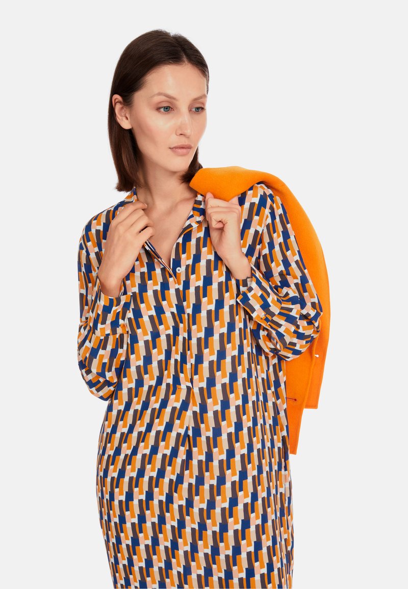 Gemustertes Kleid mit geometrischen Formen in Orange, Blau, Braun und Weiß. Leichter Stoff mit Kragen und langen Ärmeln. Orangefarbener Pullover über die Schulter drapiert.