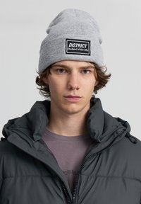 Berretto in maglia grigio con una toppa nera etichettata "DISTRICT" e la scritta "Il cuore della città"; indossato con una giacca a vento scura.
