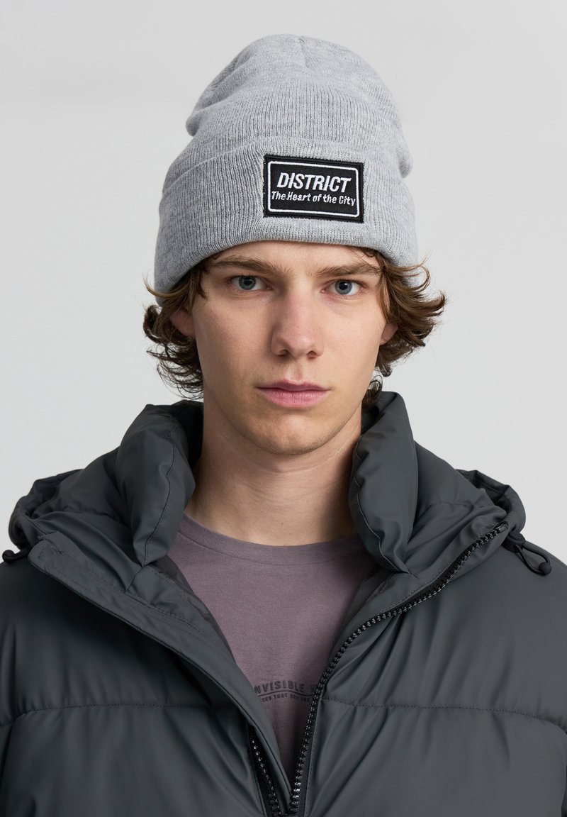 Berretto in maglia grigio con una toppa nera etichettata "DISTRICT" e la scritta "Il cuore della città"; indossato con una giacca a vento scura.