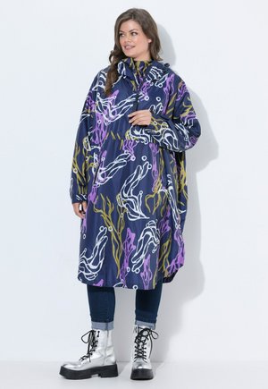 Mujer con un abrigo largo azul marino con patrones abstractos en blanco, púrpura y amarillo, vaqueros oscuros remangados y botas plateadas con cordones.