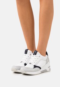 Vita och grå sportskor med texturerade material, snörning och kontrasterande marinblå detaljer. Har en vadderad häl och en modern design.