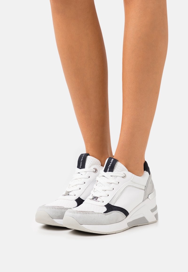 Vita och grå sportskor med texturerade material, snörning och kontrasterande marinblå detaljer. Har en vadderad häl och en modern design.