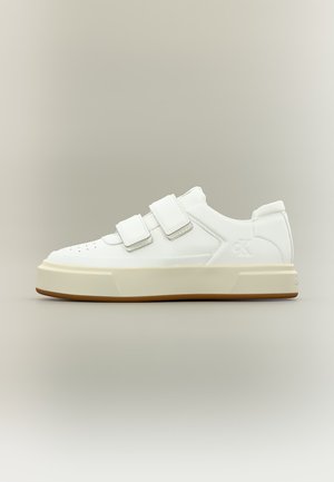 Baskets basses blanches avec deux bandes Velcro, semelle épaisse beige et logo "ck" discret sur le côté, sur un fond clair uni.