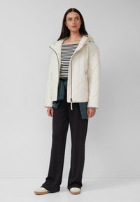 Eine Frau trägt eine weiße gesteppte Jacke über einem schwarz-weiß gestreiften Oberteil, schwarze weit geschnittene Hosen und weiße Sneakers und steht vor einer schlichten Wand.