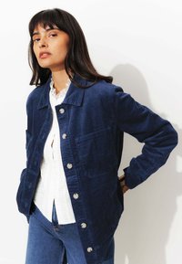Veste en velours côtelé bleu marine avec fermeture à boutons, deux poches avant et une texture lisse, portée par-dessus un chemisier blanc avec un col en dentelle.