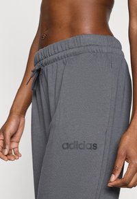 Sive bombažne športne hlače imajo elastični pas z zavezovanjem, stranske žepe in črno Adidasevo logotip na spodnjem delu noge.