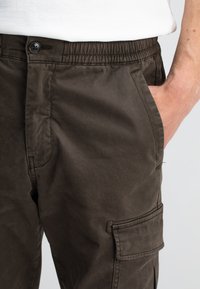 Donkerbruine cargobroek met een elastische tailleband, voorzakken en een zijzak. De stof heeft een gladde textuur en een matte afwerking.
