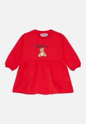 Czerwona bawełniana sukienka z długimi rękawami, okrągłym dekoltem, grafiką misia pluszowego oraz dwoma kieszeniami w kształcie serca na wysokości talii, metka marki na górze.